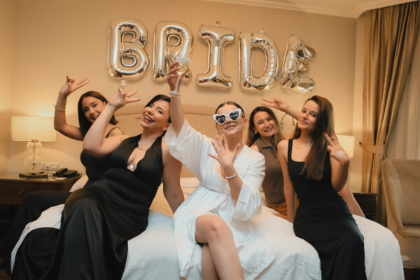 Wieczór panieński, panna młoda i jej przyjaciółki siedzą na łóżku hotelowym, z tyłu napis z balonów o treści BRIDE